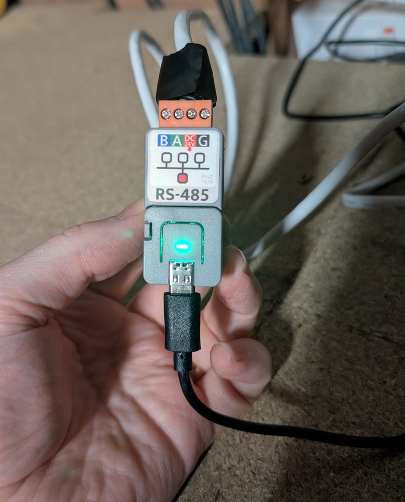 A hand holding the small grey M5Stack Atom Lite on its RS485 base, with the inverter cable taped into the screw terminals on top and its status LED glowing green.
