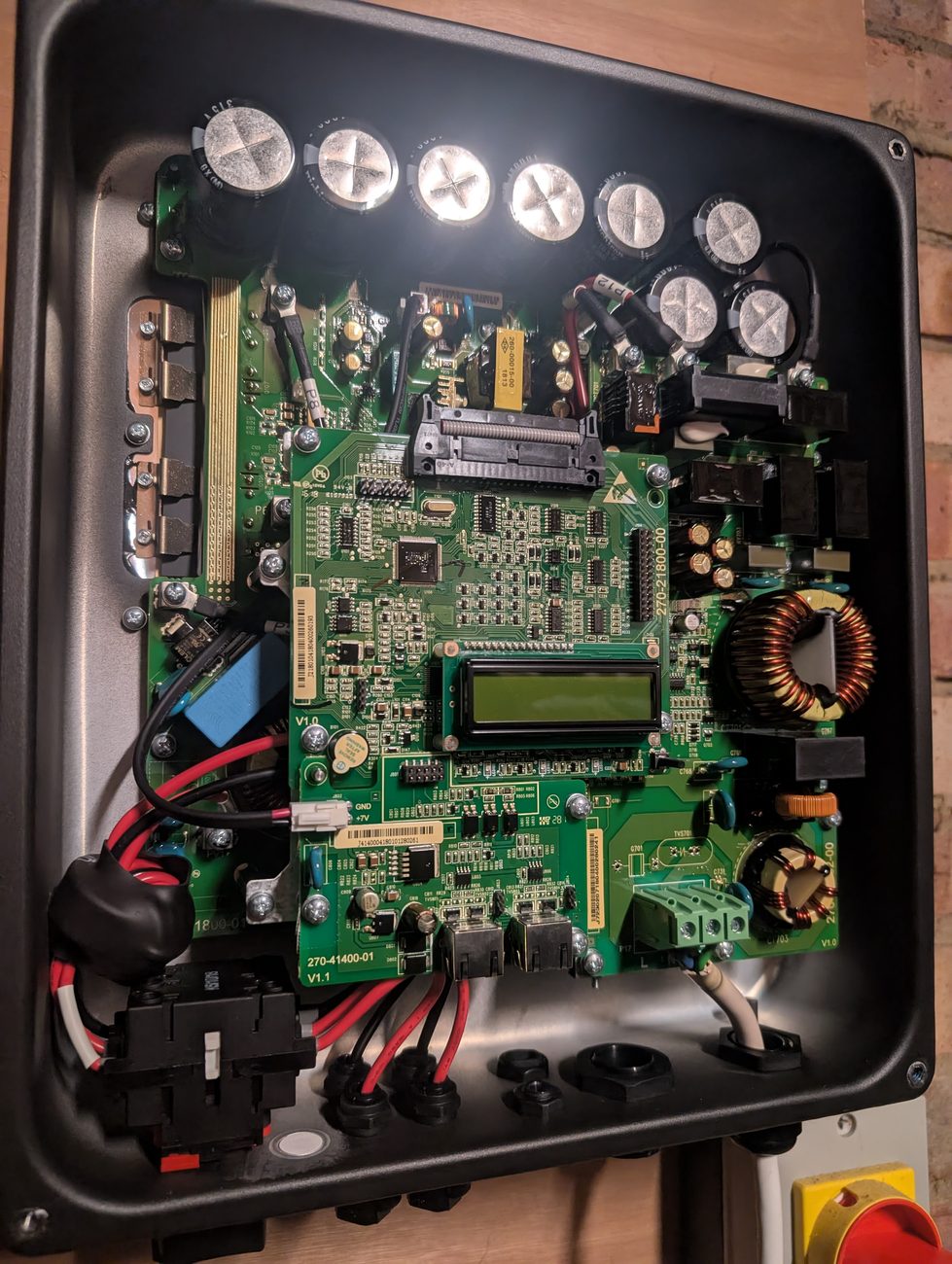 The inverter with its cover removed, showing large capacitors along the top, a green circuit board with a small LCD display, and cable glands along the bottom edge.